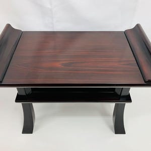 Può includere: Tavolo in legno scuro con finitura lucida. Il tavolo ha un piano rettangolare con bordi curvi e un ripiano inferiore. Le gambe nere sono curve. Il tavolo è di colore marrone rossiccio scuro.