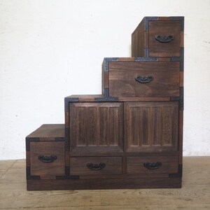 May include: A dark brown wooden chest of drawers with five drawers and a stepped design. The drawers have black metal handles and the chest has black metal accents.