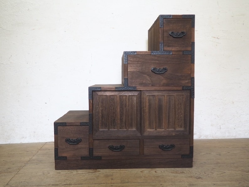 May include: A dark brown wooden chest of drawers with five drawers and a stepped design. The drawers have black metal handles and the chest has black metal accents.