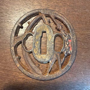 Op de afbeelding: Een cirkelvormige Japanse tsuba, of zwaardbeschermer, met een centrale opening en een ingewikkeld opengewerkt ontwerp. Het metaal heeft een verweerde, roestbruine uitstraling met gouden en rode accenten, wat duidt op leeftijd en gebruik.