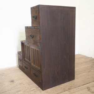 May include: A dark brown wooden chest of drawers with multiple tiers. The chest has a traditional Japanese design with a slatted front panel and metal accents.