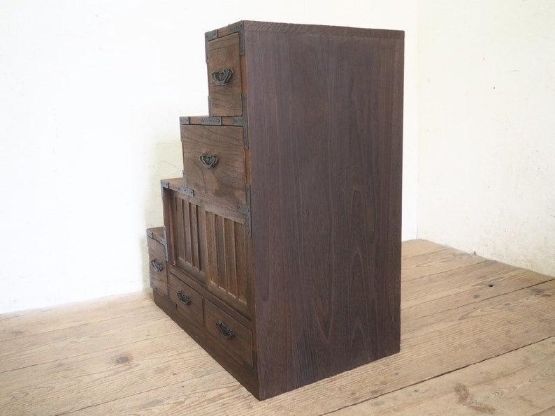 May include: A dark brown wooden chest of drawers with multiple tiers. The chest has a traditional Japanese design with a slatted front panel and metal accents.