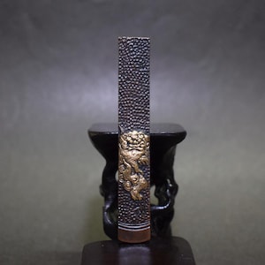 May include: A dark brown, rectangular object with a textured surface and a gold-coloured decorative element depicting a mythical creature. The object stands upright on a dark surface. The gold element is near the bottom of the object.