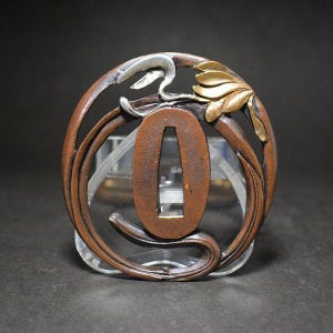 Op de afbeelding: Een antieke Japanse tsuba, een zwaardbeschermer, met een ovale vorm en ingewikkelde bloemen- en wijnstokontwerpen. De beschermer is voornamelijk van brons met zilveren en gouden accenten, wat gedetailleerd vakmanschap en een rijke, verouderde patina laat zien.