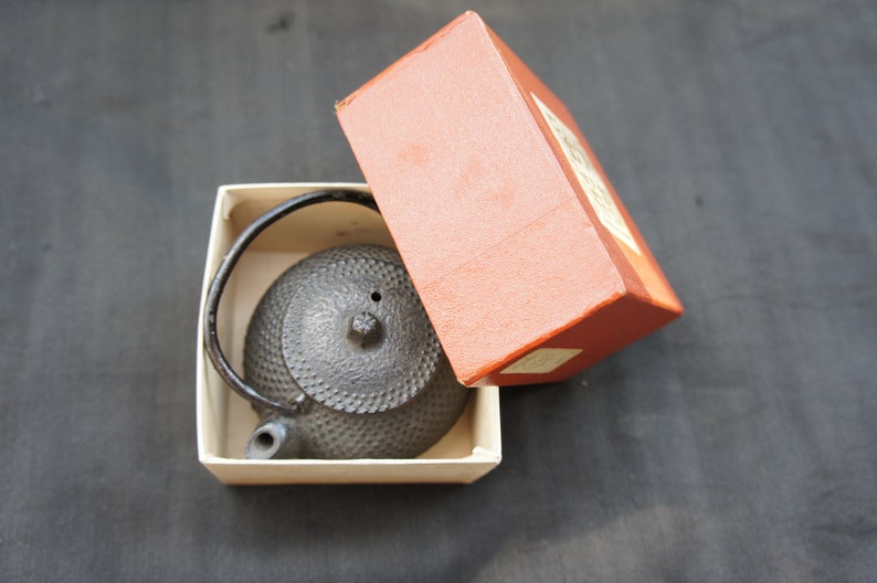 Op de afbeelding: Een zwarte gietijzeren theepot met een getextureerd oppervlak zit in een witte doos met een rood deksel. Het deksel heeft Japanse tekst.