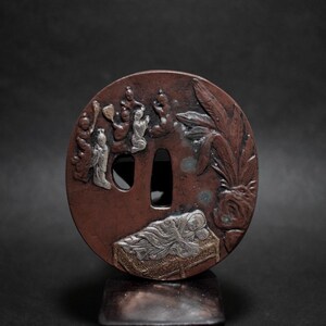 Op de afbeelding: Een ronde, roodbruine Japanse tsuba van metaal met ingewikkelde reliëfsnijwerk. Het ontwerp toont figuren, een slapende persoon en bladeren. De tsuba heeft twee openingen en is waarschijnlijk een decoratief zwaardonderdeel.