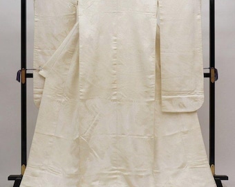 May include: A cream-colored Japanese kimono with a subtle floral pattern, displayed on a black stand. The kimono features long sleeves and a floor-length design, showcasing traditional Japanese attire.