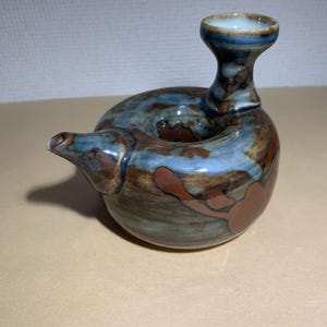 May include: A handcrafted ceramic teapot with a unique, organic shape. The teapot features a spout, a handle, and a small opening at the top. The color palette includes shades of blue, brown, and cream, with a glossy finish. The teapot is a decorative item.