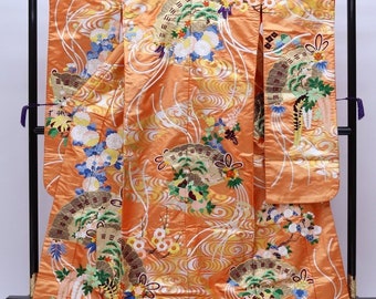 May include: An orange kimono with intricate embroidery of fans, flowers, and swirling patterns. The traditional Japanese garment is displayed on a black stand, showcasing its detailed design and vibrant colors. The kimono's design includes white, blue, and green accents.