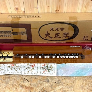 以下が含まれることがあります： 段ボール箱に入ったスズキの大正琴。楽器は磨かれた茶色の木製ボディ、金色のペグ、白い番号付きのキーが並んでいます。箱には「スズキ」という文字と日本語の漢字が印刷されています。