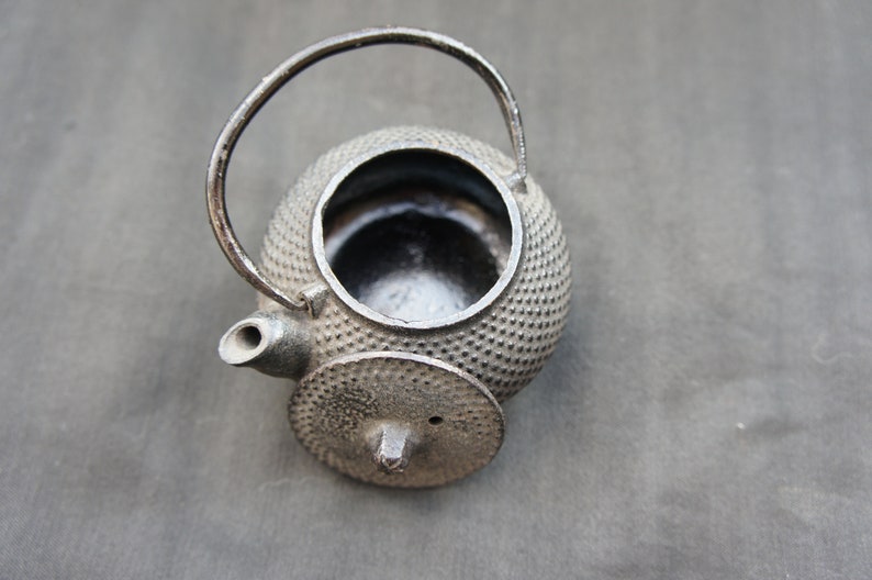 Op de afbeelding: Een zwarte gietijzeren theepot met een rond lichaam en een kleine tuit. De theepot heeft een getextureerd oppervlak en een deksel met een klein gaatje in het midden.