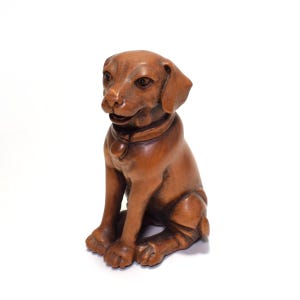 May include: A wooden sculpture of a seated dog with a collar and a pendant. The dog is carved from a light brown wood and has detailed features, including its eyes, nose, and ears. The sculpture is on a white background.