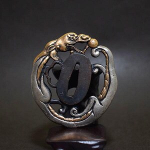 Op de afbeelding: Een close-up van een Japanse katana tsuba, een handbeschermer. De tsuba heeft een drakenontwerp in goud- en zilverkleuren, met ingewikkelde details. De centrale opening is donker en de algehele esthetiek is sierlijk en historisch.