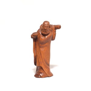 Pode incluir: Escultura em madeira de uma figura vestida com uma túnica, com uma longa barba, carregando um bastão sobre o ombro. A escultura é esculpida em madeira castanha clara e ergue-se sobre um fundo branco. Os traços faciais da figura são detalhados.