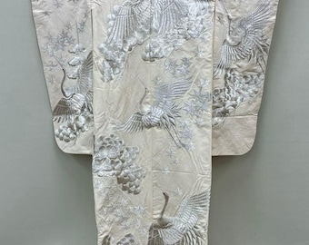 May include: Cream-colored kimono with intricate silver embroidery of cranes, clouds, and stars. The kimono has a red trim along the bottom and sleeves. The design is symmetrical, with the cranes appearing to be in flight.