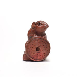 May include: A carved wooden sculpture of a brown rat holding a large, round coin. The coin has a square hole in the center and is covered in small, evenly spaced holes. The rat is detailed with visible ears and eyes.