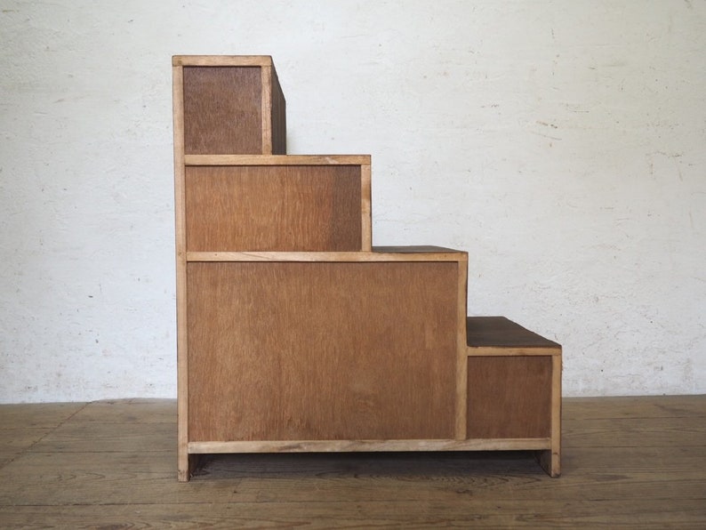 May include: A wooden step shelf with four tiers, each with a separate compartment. The shelf is made of light brown wood and has a simple, minimalist design.
