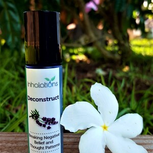 Pode incluir: Um frasco preto com rolo com um rótulo branco que diz "Inhalations Deconstruct Breaking Negative Belief and Thought Patterns 35 oz". O frasco está sobre uma superfície de madeira com uma flor branca.