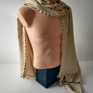 May include: A beige scarf with dark brown stitching details, draped over a mannequin. The scarf has a textured appearance and a fringed edge. The mannequin is on a dark blue base, against a white background. The scarf appears to be made of a soft, natural fabric.