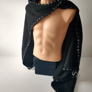 May include: Black hooded cape with white stitching detail. The cape is displayed on a mannequin and features a circular clasp closure. The edges are finished with white stitching. The cape is made of a soft, textured fabric.