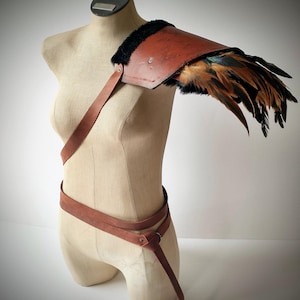 May include: A brown leather shoulder pauldron with black fur trim and attached feathers. The pauldron is part of a costume and is displayed on a mannequin. A brown leather belt with a strap is also included.