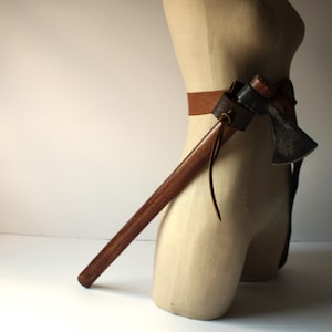 May include: A brown leather belt with a brown leather sheath for a small axe. The axe has a brown wooden handle and a metal head. The axe is attached to the belt and is hanging down.