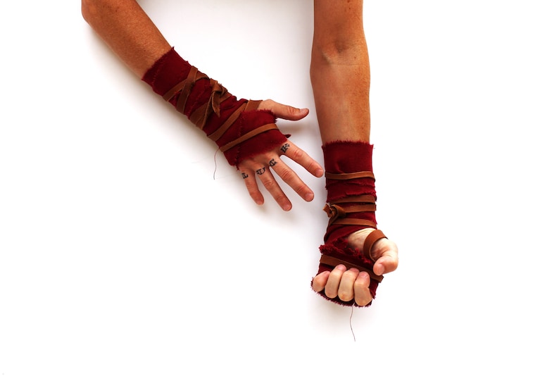 Puede incluir: Un par de mu&ntilde;equeras de tela roja con correas de cuero marr&oacute;n. Las mu&ntilde;equeras est&aacute;n atadas alrededor de las mu&ntilde;ecas y aseguradas con nudos.