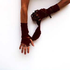 Puede incluir: Los brazos de una persona est&aacute;n envueltos en tela roja con correas de cuero marr&oacute;n. La tela est&aacute; rasgada y deshilachada. Las manos de la persona son visibles, con tatuajes de tinta negra en los dedos.