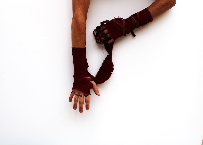 Puede incluir: Los brazos de una persona est&aacute;n envueltos en tela roja con correas de cuero marr&oacute;n. La tela est&aacute; rasgada y deshilachada. Las manos de la persona son visibles, con tatuajes de tinta negra en los dedos.