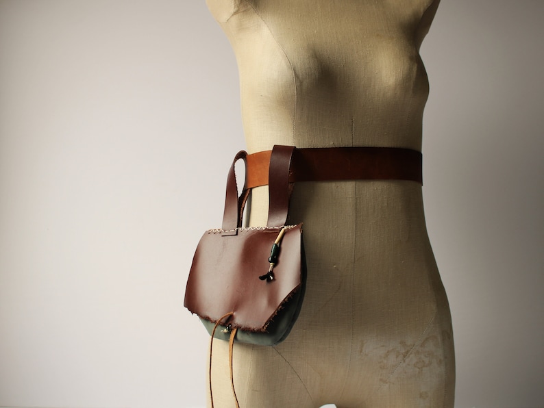 May include: A brown leather bag with a flap and a drawstring closure. The bag is attached to a brown leather belt.