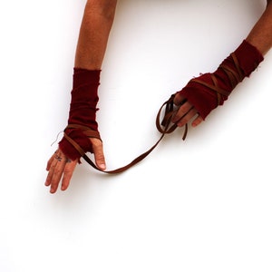 Puede incluir: Un par de calentadores de brazo de tela roja con correas de cuero marr&oacute;n envueltas alrededor. Las correas est&aacute;n atadas juntas en la mu&ntilde;eca.