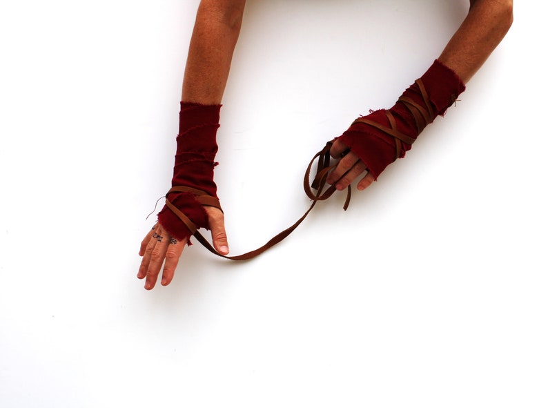 Puede incluir: Un par de calentadores de brazo de tela roja con correas de cuero marr&oacute;n envueltas alrededor. Las correas est&aacute;n atadas juntas en la mu&ntilde;eca.