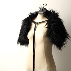 May include: Black faux fur shrug with a black strap. The shrug is displayed on a white mannequin.