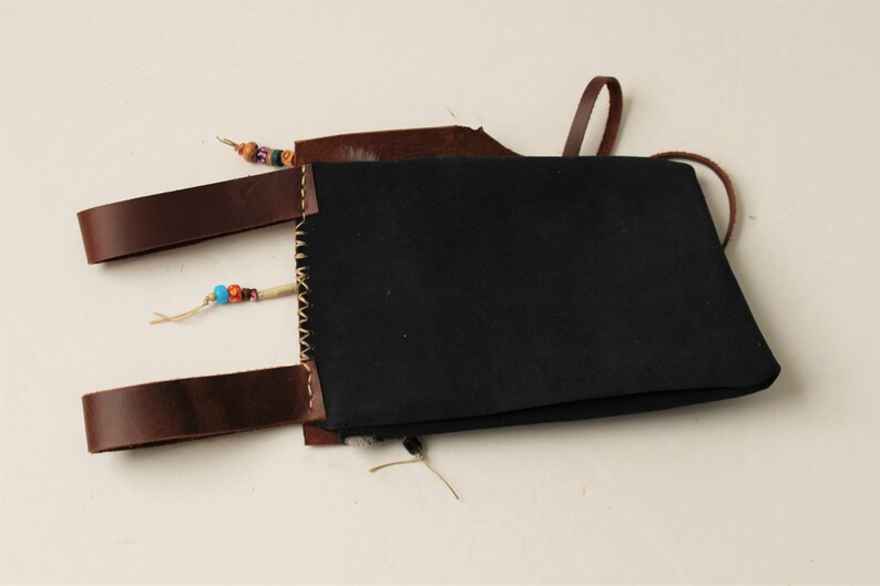 Puede incluir: Una bolsa de cuero negro con correas de cuero marr&oacute;n y detalles de cuentas de colores. La bolsa tiene un cierre de solapa y una correa larga para llevar.