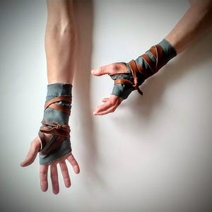 May include: A pair of handmade arm wraps. The wraps are made of gray fabric and brown leather straps. The leather straps are wrapped around the fabric and tied in place. The arm wraps are designed to protect the forearm and wrist.