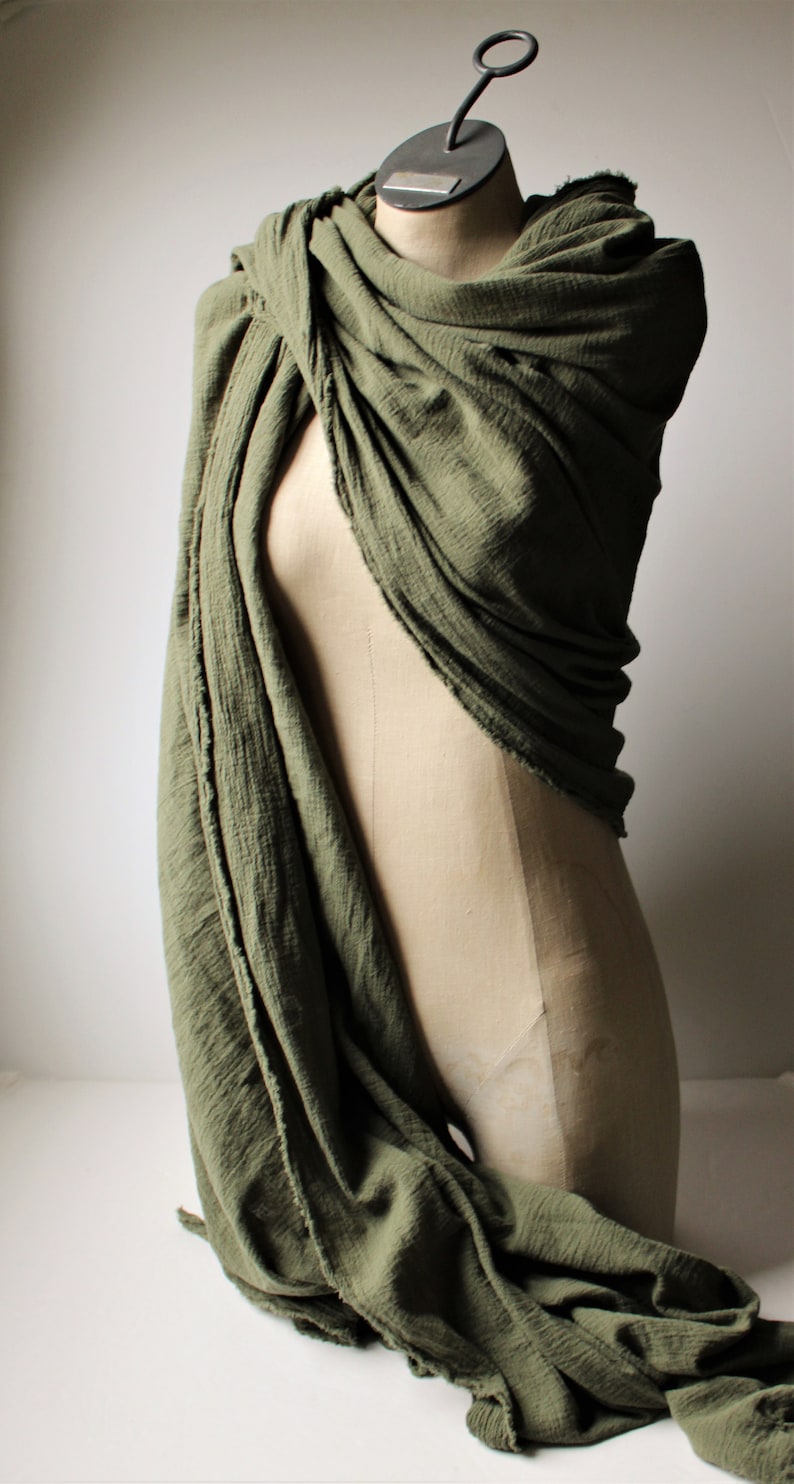 May include: Olive green scarf draped over a mannequin. The fabric has a textured appearance and is loosely wrapped, creating soft folds. The mannequin has a metal hanger attached to the top.