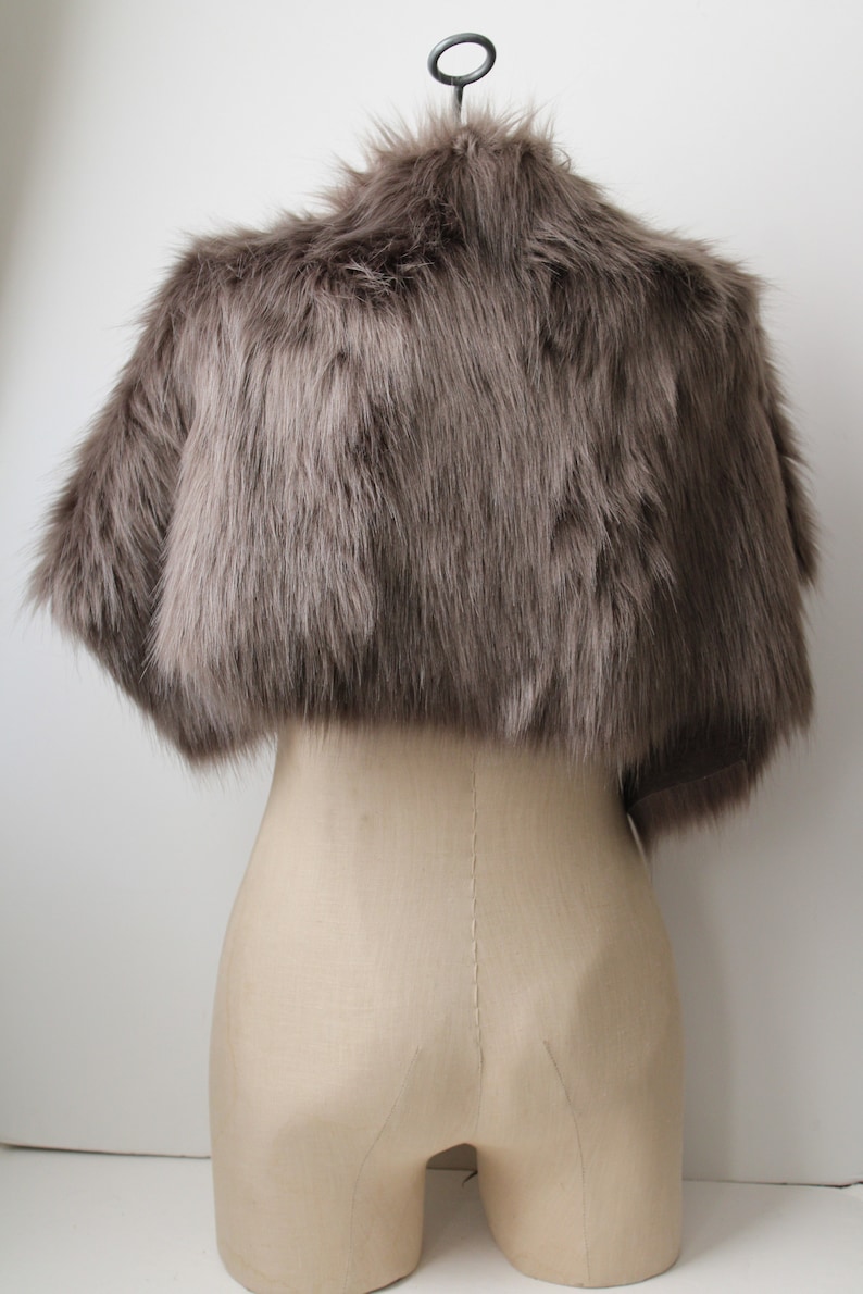 May include: A gray faux fur shrug with a short, cropped length. The shrug is displayed on a mannequin against a white background.
