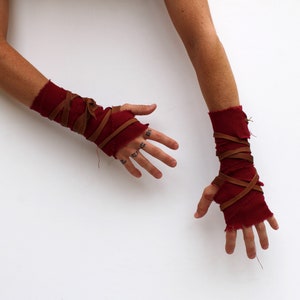 Puede incluir: Un par de calentadores de brazo de tela roja con correas de cuero marr&oacute;n envueltas alrededor. Los calentadores de brazo se usan en ambos brazos y las manos son visibles.