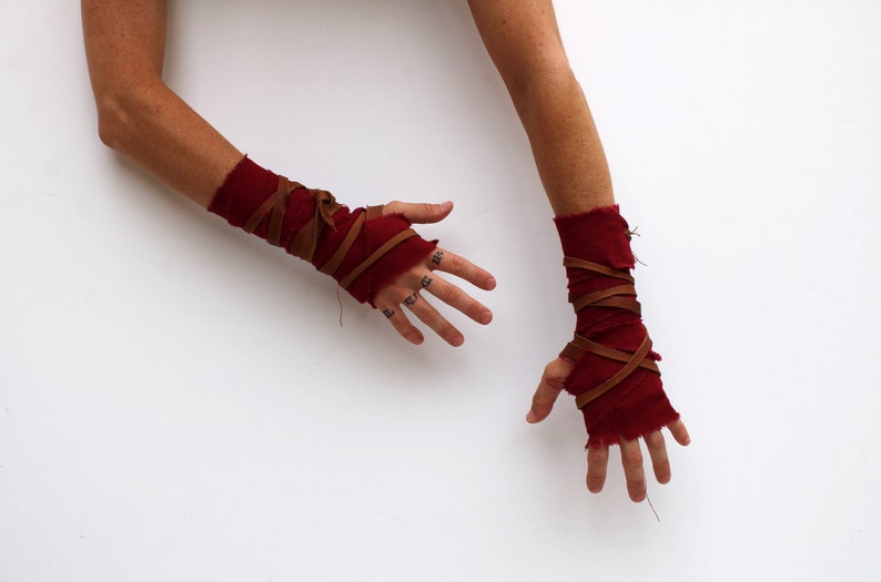 Puede incluir: Un par de calentadores de brazo de tela roja con correas de cuero marr&oacute;n envueltas alrededor. Los calentadores de brazo se usan en ambos brazos y las manos son visibles.