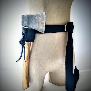 May include: A black leather belt with a silver buckle holds a small axe in a black leather sheath. The axe has a wooden handle and a silver head. The belt is displayed on a mannequin.