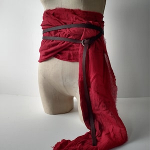 May include: A red fabric wrap belt with a dark brown leather belt wrapped around it. The belt is draped around a mannequin. The fabric has a frayed edge. The leather belt has a metal ring closure.