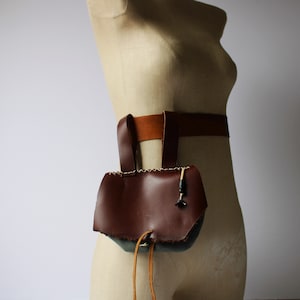 May include: A brown leather pouch with a flap and a drawstring closure. The pouch is attached to a brown leather belt. This is a renaissance style belt bag for a costume.