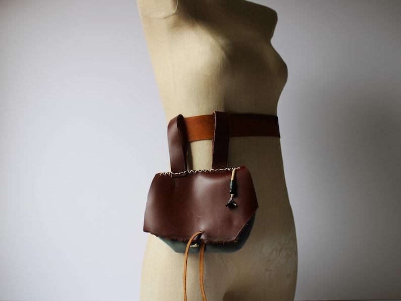 May include: A brown leather pouch with a flap and a drawstring closure. The pouch is attached to a brown leather belt. This is a renaissance style belt bag for a costume.