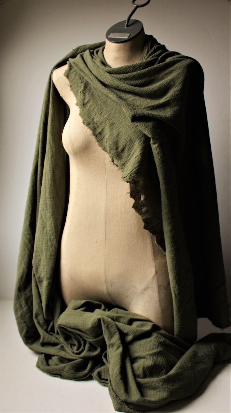 May include: Olive green scarf draped over a mannequin. The fabric has a textured appearance and a frayed edge. The scarf is wrapped around the neck and cascades down the front of the mannequin, creating soft folds.