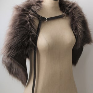 May include: A gray faux fur shrug with a gray leather strap with silver hardware. The shrug is displayed on a mannequin.
