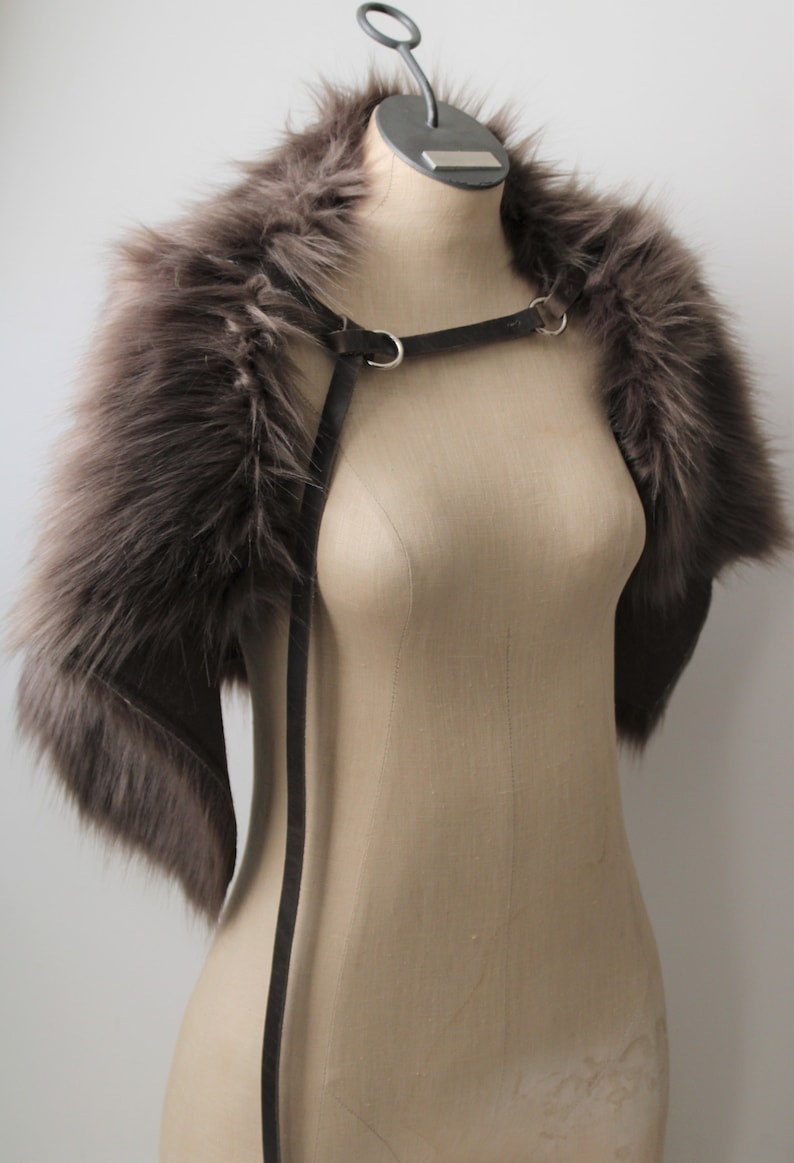 May include: A gray faux fur shrug with a gray leather strap with silver hardware. The shrug is displayed on a mannequin.