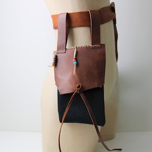 May include: A brown leather belt with a brown leather pouch attached. The pouch has a drawstring closure. The belt has a decorative beaded strap with colorful beads.