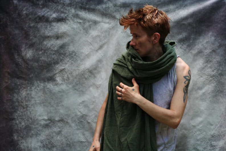 May include: A person wearing a gray tank top and a green scarf. The person has short red hair and tattoos on their arm and hand. The background is a textured gray.