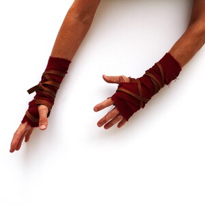 Puede incluir: Un par de calentadores de brazo de tela roja con correas de cuero marr&oacute;n envueltas alrededor. Los calentadores de brazo se usan en ambos brazos y las manos est&aacute;n extendidas.
