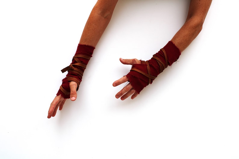 Puede incluir: Un par de calentadores de brazo de tela roja con correas de cuero marr&oacute;n envueltas alrededor. Los calentadores de brazo se usan en ambos brazos y las manos est&aacute;n extendidas.